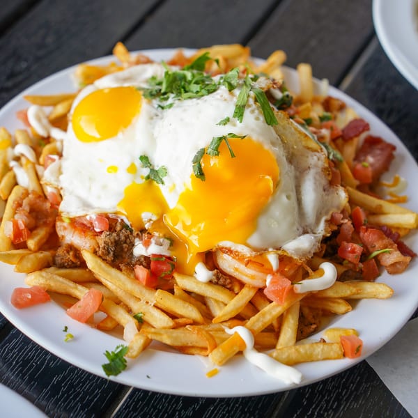Loaded fries topped with two fried eggs, bacon, tomatoes, and cilantro on a white plate.