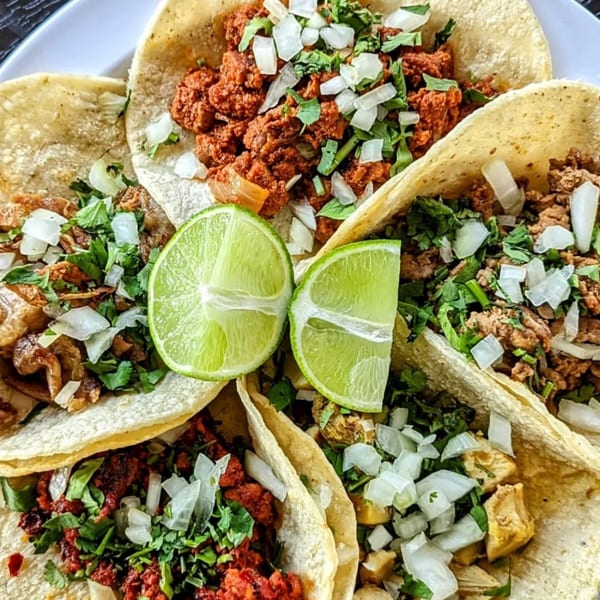A plate of five street tacos with various fillings, cilantro, onions, and two lime wedges.