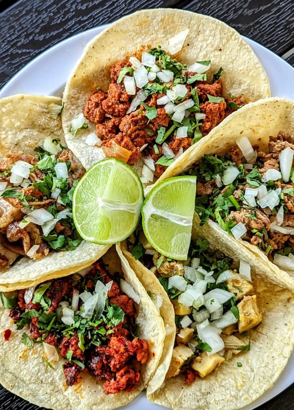 A plate of five street tacos with various fillings, cilantro, onions, and two lime wedges.