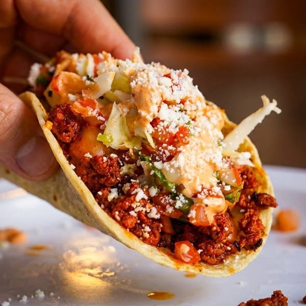 A hand holds a delicious taco filled with meat, lettuce, cheese, and sauce over a white plate.