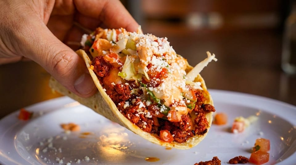 A hand holds a delicious taco filled with meat, lettuce, cheese, and sauce over a white plate.