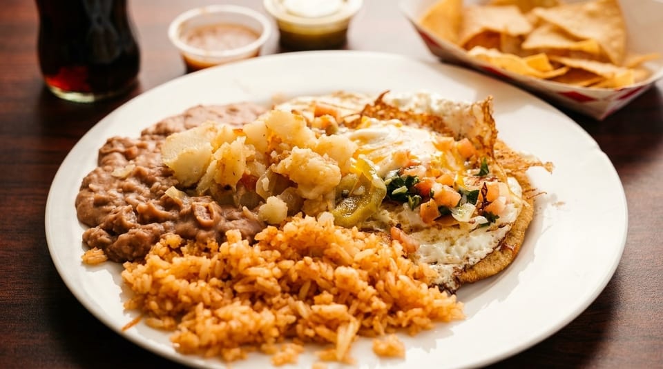 A plate of huevos rancheros with rice, refried beans, and potatoes, with chips and a drink.